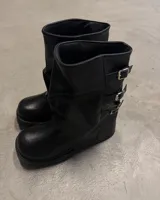 Bota corta negra de estilo motociclista, caña arrugada y suela plataforma gruesa. Presenta tres hebillas metálicas ajustables en el lateral exterior.