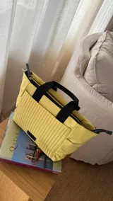 Cartera tipo tote de nylon color malva, con diseño capitoneado de líneas verticales en el frente y rombos en los laterales. Cuenta con doble asa de mano en color negro, bolsillos laterales y un parche decorativo con logo en la parte frontal.