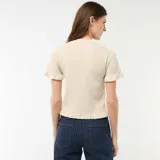 Camiseta de punto color beige con cuello redondo, cierre frontal de botones y mangas cortas con volantes. Presenta detalles de volantes en la botonera y en el ruedo.