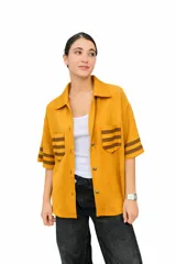 Camisa de algodón color mostaza, con cuello solapa, mangas cortas con rayas marrones y bolsillos frontales con solapa y rayas marrones. Cierre frontal con botones.