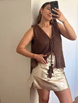 Chaleco tejido en punto calado con hilo de algodón y lurex, de color negro con destellos plateados. Presenta escote profundo en V, cierre frontal con cordones entrelazados terminados en borlas y un corte recto y holgado.