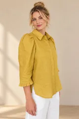 Camisa de lino color blanca, con cuello clásico, mangas tres cuartos abullonadas en los hombros y terminación con volados en los puños, cierre frontal con botones ocultos.
