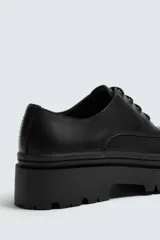 Zapato de vestir tipo derby, color negro, con parte superior lisa, horma redonda, suela con plataforma y piso track.