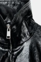 Chaqueta de efecto piel en color negro con cuello subido y bufanda desmontable integrada mediante botones. Presenta manga larga con trabillas ajustables en los puños, cierre frontal con cremallera y bolsillos laterales ocultos en las costuras. El diseño incluye un bajo con elástico en la espalda y forro interior.