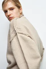 Jersey de punto color beige con cuello subido, manga larga y detalle de botones en los puños. Presenta costuras marcadas y acabados en rib.