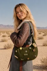 Cartera tipo baguette de gamuza color verde oliva, adornada con diversos apliques metálicos, botones y dijes decorativos. Cuenta con una placa metálica con la marca Agnes en el frente y una correa de hombro integrada.