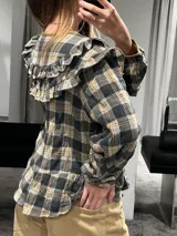 Camisa de manga larga con estampado a cuadros en tonos gris, negro y mostaza. Presenta volados en la parte superior delantera y en los hombros, con un diseño de corte holgado y lazos decorativos en el cuello.