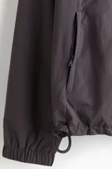 Campera rompevientos de nylon repelente al agua, color gris oscuro/marrón topo, con forro de malla. Presenta cierre frontal con protección para el mentón, capucha ajustable con cordón, bolsillos laterales y bolsillo interior con cierre, y elástico revestido en puños y bajo.
