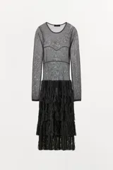 Vestido midi negro, semitransparente, de manga larga y cuello redondo. La parte superior presenta un tejido de punto fino con costuras que simulan un bustier. La falda está compuesta por capas de flecos largos y densos.
