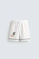 Bermuda de corte relajado confeccionada en algodón, color blanco con vivo en contraste color marrón en el ruedo. Presenta cintura elástica con cordón ajustable y un bordado decorativo en la pierna derecha que incluye una palmera y un gallo.