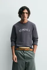 Camiseta de manga larga, corte ajustado (muscle fit), color gris oscuro con efecto lavado. Presenta un estampado frontal desgastado con el texto curvo "ST MONICA" en color plateado o gris claro.