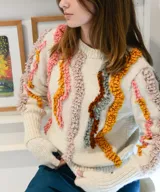 Sweater tejido a mano en punto jersey de 100% lana merino, color blanco con detalles texturizados en punto bucle de tonos rosados, mostaza, marrón y gris, formando líneas onduladas en el frente y las mangas. Cuenta con cuello redondo base y puños largos acANALados.