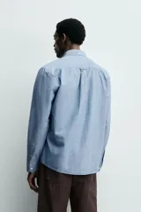 Camisa de corte regular fit confeccionada en tejido chambray de algodón con efecto lavado. Presenta cuello con botones, manga larga con puños abotonados, bolsillo en el pecho y cierre frontal de botones.