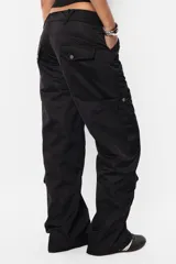 Pantalón cargo negro de corte recto y tiro bajo, con recortes y múltiples bolsillos laterales y traseros con tapa y botón a presión. Cierre frontal con cremallera de nylon y botón personalizado.