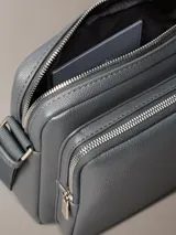 Bolso bandolera Calvin Klein color gris oscuro, de cuero sintético con textura granulada. Tiene correa ajustable, cierre superior con cremallera, bolsillo frontal con cremallera y logo de la marca grabado en la parte inferior.