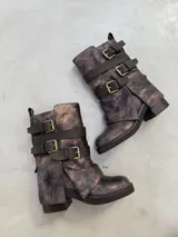Botas de caña media en tono marrón con efecto metalizado y textura desgastada. Cuentan con múltiples tiras horizontales con hebillas metálicas doradas y cierre lateral. Poseen taco ancho y suela track.