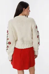 Cárdigan tejido a mano color crudo, con diseño de trenzas y flores bordadas en azul y marrón. Tiene cuello redondo y cierre frontal con botones de madera.