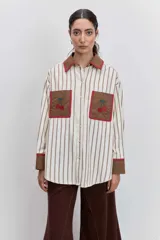 Camisa de corte oversize con estampado de rayas verticales, cuello clásico y cierre frontal de botones. Presenta dos bolsillos frontales en contraste con bordados de tréboles, además de puños y cuello en color sólido con detalles en amarillo.