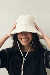 Gorro tipo bucket de color blanco crudo, con borde desflecado y cordón ajustable con tope.