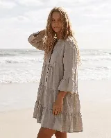 Vestido corto de lino natural color gris, con diseño amplio de capas, escote en V con botones frontales y mangas largas con volados en los puños.
