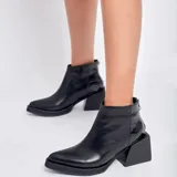 Botín de cuero vacuno color negro, con diseño combinado de cuero liso en la parte delantera y cuero con textura de reptil en el talón. Cuenta con un taco cuadrado de madera, cierre metálico en la parte posterior y suela de goma.