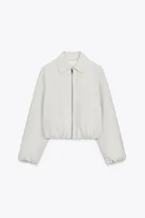 Chaqueta bomber corta color blanco hueso, con cuello solapa, manga larga y falsos bolsillos delanteros. Presenta acabados abullonados y elásticos en puños y bajo, y cierre frontal con cremallera.