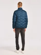 Chaqueta acolchada de peso medio en color azul petróleo, con cierre frontal, cuello alto y parche de silicona con logo en el pecho. Cuenta con bolsillos laterales y ribetes elásticos en puños y dobladillo.