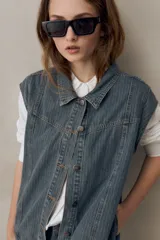 Chaleco de jean azul con sutil textura a rayas, cuello camisero, cierre frontal con botones metálicos y diseño sin mangas con hombros amplios.