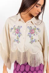 Camisa corta de color beige con silueta amplia, mangas anchas tipo capa terminadas en flecos y ramos de flores bordados en el frente.