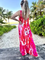 Vestido largo estampado en tonos vibrantes de fucsia, rosa, naranja y blanco, con diseño abstracto y palmeras. Presenta tirantes anchos y escote pronunciado en la espalda, con un ruedo con volado.