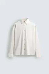 Camisa blanca de algodón con corte relajado, cuello solapa, manga larga con puño abotonado y bolsillo de plastrón en el pecho. Presenta bajo asimétrico con aberturas laterales y cierre frontal de botonadura.
