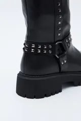 Bota de caña media estilo biker, confeccionada en cuero sintético color marrón oscuro. Presenta una suela track robusta, detalle de tachas metálicas verticales a lo largo de la caña y un arnés decorativo con argolla metálica en el tobillo.