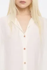 Camisa larga color blanco, de corte oversize y escote con solapas. Tiene manga corta recta y ancha, cierre con botonadura frontal y bajo ajustable con cordones de tejido.