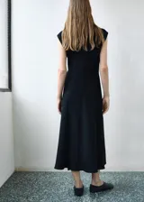 Vestido midi de color negro, sin mangas, con cuello redondo y diseño entallado. Presenta un detalle de fruncido lateral y cierre de cremallera en la parte posterior.