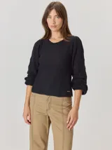 Buzo de lana color beige con mangas abullonadas de tejido calado con diseño de rombos. Presenta un cuello redondo y un detalle de flor negra de tela con cintas colgantes en el escote.