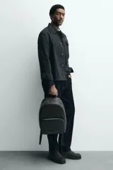 Mochila negra con textura grabada, compartimento principal con cierre de cremallera, bolsillo interior con cremallera, bolsillo frontal con cremallera, parte posterior acolchada con banda de sujeción para equipaje, asa de mano y dos asas de hombro ajustables.