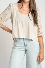 Blusa color arena de algodón peinado, con escote redondo, mangas cortas abullonadas con bordados suizos y detalles de alforcas en el frente.