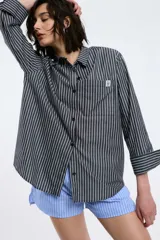 Camisa de popelina rayada en color azul marino y blanco, con corte oversized, abotonadura frontal y en las mangas, y un bolsillo frontal con un parche blanco.
