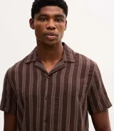 Camisa de manga corta con cuello solapa y cierre frontal de botones. Presenta un diseño de rayas verticales con textura jacquard en tonos marrones.
