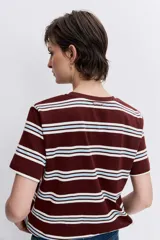 Remera de manga corta con cuello redondo, confeccionada en tejido de punto con diseño de rayas horizontales en tonos marrón, blanco y celeste. Presenta un pequeño parche circular rojo en el pecho.