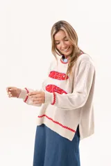 Sweater tejido color crudo con cuello redondo y mangas largas. Presenta dos peces rojos con detalles en azul bordados en el frente. Tiene ribete ondulado rojo en cuello, puños y ruedo.