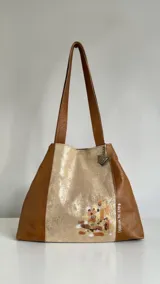 Cartera tipo shopper de cuero, con diseño combinado en colores caramelo y perla, asas de hombro y arte pintado a mano en el frente.