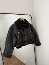 Campera tipo aviador de cuero sintético negro, totalmente forrada en corderito sintético al tono. Presenta cuello con solapas, cierre frontal con cremallera, bolsillos laterales con cierre y cinturón ajustable en el ruedo con hebilla metálica.