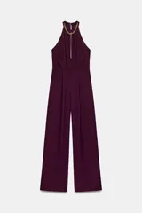 Mono largo de color bordó con cuello halter adornado con una cadena dorada y un pronunciado escote en V con abertura central. Presenta pinzas en la cintura y pernera ancha, con cierre trasero de cremallera oculta y botones.