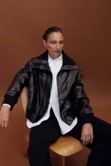 Chaqueta de cuero estilo bomber en color negro, con cuello alto, cierre frontal con cremallera y solapa con botones a presión. Presenta puños y cintura de tejido elástico acanalado, bolsillos laterales y un diseño estructurado.