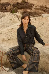 Camisa negra holgada de manga larga, con cuello clásico, botones delanteros y detalle de flor aplicada a tono en el pecho. Presenta diseño más largo en la parte trasera.