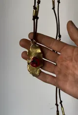 Collar artesanal con dije de base metálica irregular y piedra central roja. Presenta una estructura de tiento de gamuza marrón con detalles de tubos metálicos dorados.