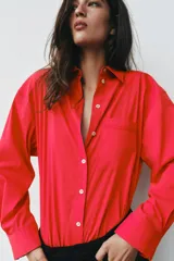 Camisa de popelina roja, de corte oversize, con cuello solapa, manga larga y bajo asimétrico redondeado. Cierre frontal oculto con botones.
