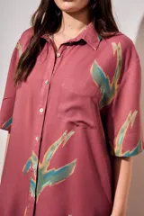 Vestido camisero corto de color rosado con estampado de aves abstractas en tonos verdes y azules. Cuenta con cuello camisero, cierre frontal con botones, mangas cortas y un bolsillo aplicado en el pecho.