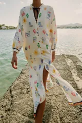 Vestido largo estilo túnica de color crudo con estampado multicolor de motivos marinos y mediterráneos. Presenta cuello con volante, cierre frontal con botones, manga larga y cordón ajustable en el cuello con abalorios.